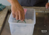 A man's hand placing a jade bangle into a plastic container filled with hot soapy water.